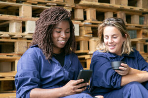 Happy African American and Caucasian factory colleagues in overalls watching content on cellphone together 